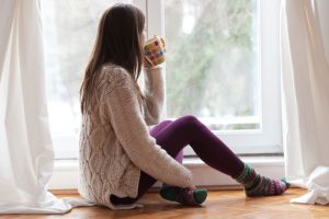 young woman dressed for cold weather sitting by the window having hot drink