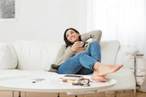 Young smiling woman sitting on sofa and looking up while drinking hot tea. Young brunette woman thinking at home in a leisure time. Happy girl relaxing at home on a bright winter morning.