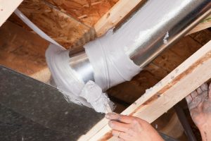 A dirty male hand, holding a paintbrush, is spreading caulk over a house air duct joint with a paintbrush. The caulk has been applied from a tube with a caulk gun and the brush is evenly spreading it over the joint. Caulking residential ducts prevents air-conditioned or heated air from escaping the duct and prevents ambient air from entering the system. This is a new practice in homebuilding to increase energy efficiency, eventually it may be come standard or mandatory.