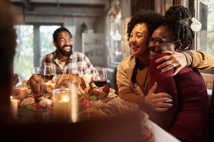 Happy African American mature mother embracing her adult daughter while having Thanksgiving lunch with adult son at dining table.