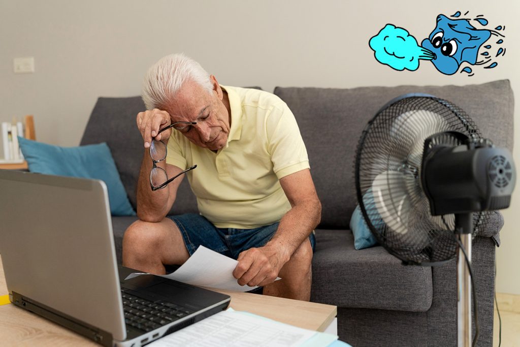 Worried senior man calculating domestic bills at home, using laptop and cooling off with a fan.