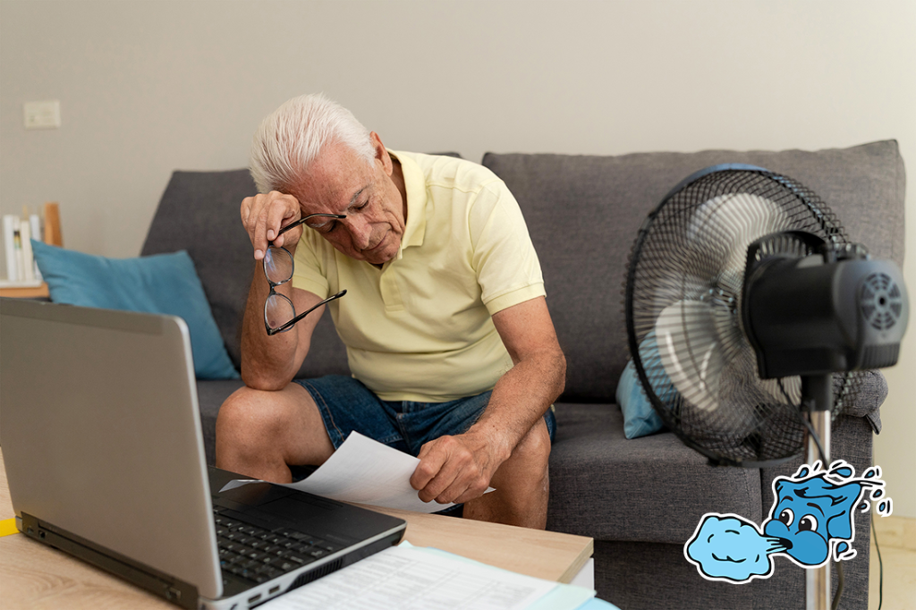 Worried senior man calculating domestic bills at home, using laptop and cooling off with a fan.