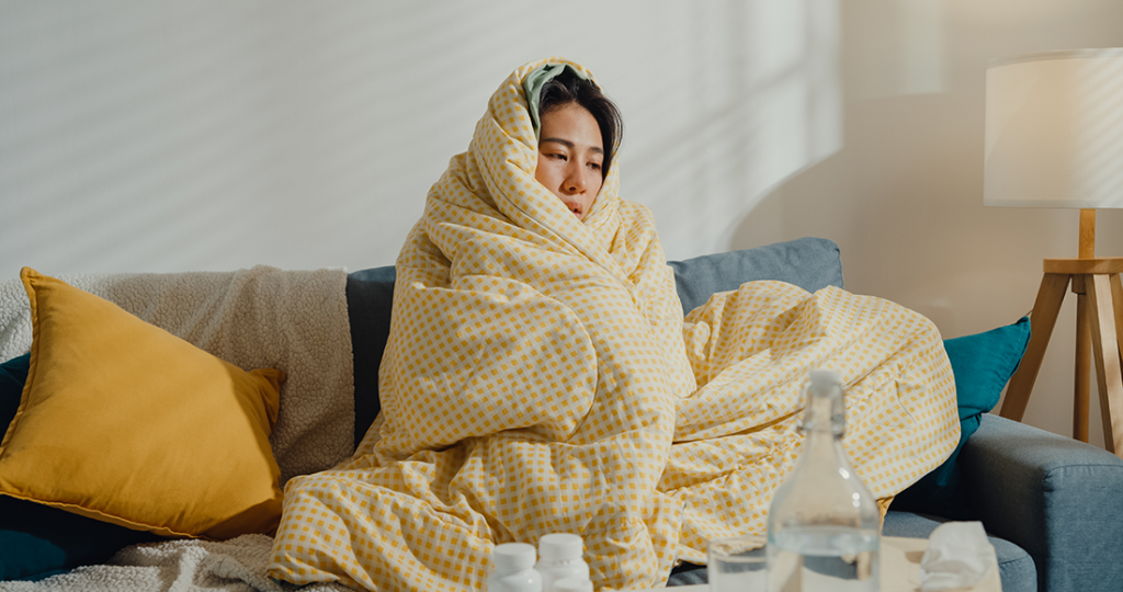 Drowsy looking woman wrapped in a yellow duvet looking miserable. There's a tissue box and a bottle of water on the coffee table in front of her.
