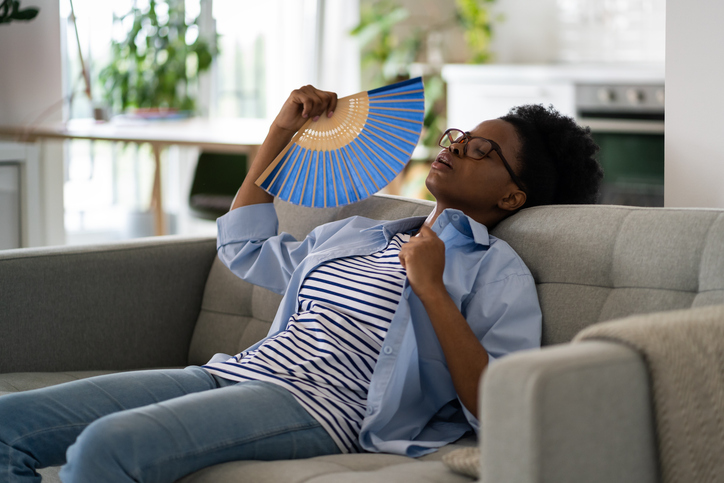 Tired exhausted African American woman sits on sofa at home using paper fans and closing eyes suffering from heat
